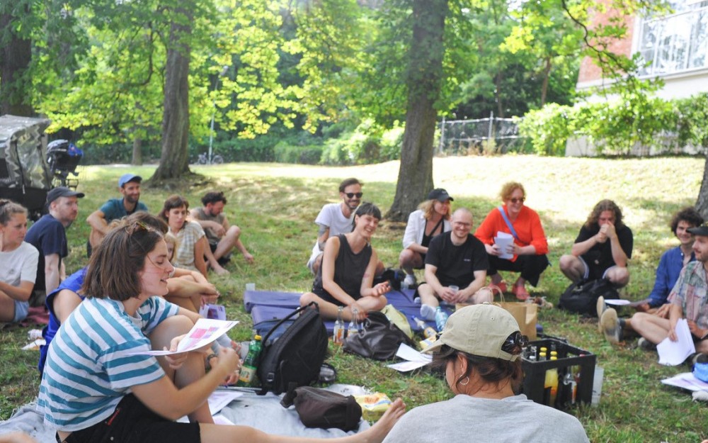 Eine Gruppe Menschen, Mitglieder des Kollektiv Kaorle,
                                                sitzt vor dem Pavillon 21 in der Wiese und bespricht wie es weiter geht.