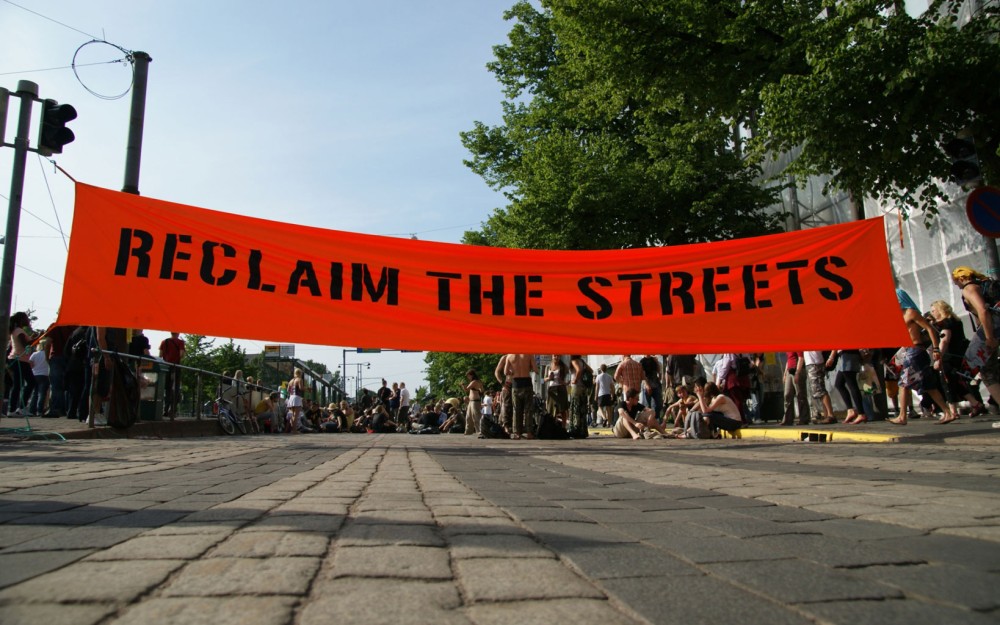 Das Bild
                                                zeigt eine gepflasterte Straße, über die ein roter Banner mit dem Schriftzug "Reclaim the streets" gespannt ist.