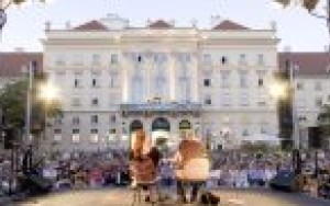 Im Vordergrund die Bühne von hinten, auf der Bühne sitzen 2 Personen, im Hintergrund die Fassade des Hauptgebäudes
                                 vom Museumsquartiers, dazwischen zahlreiche Menschen, die der Veranstaltung beiwohnen.